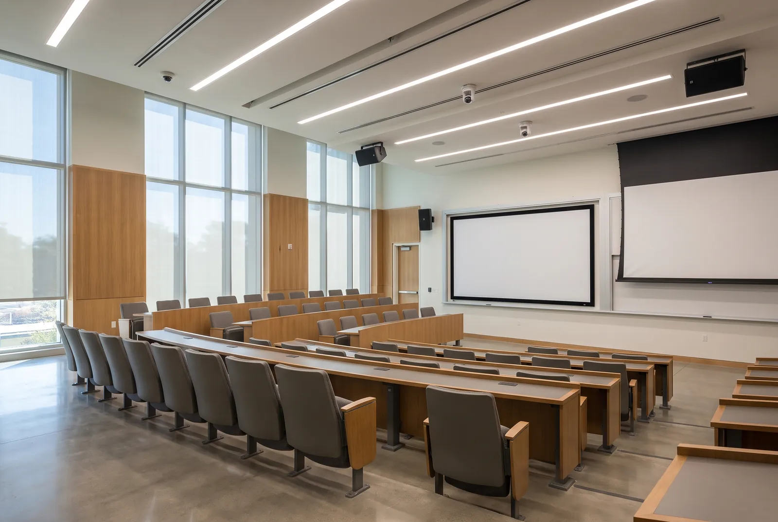 A tiered lecture room with rows of desks and seats, two projection screens, ceiling speakers and tall windows along one side.