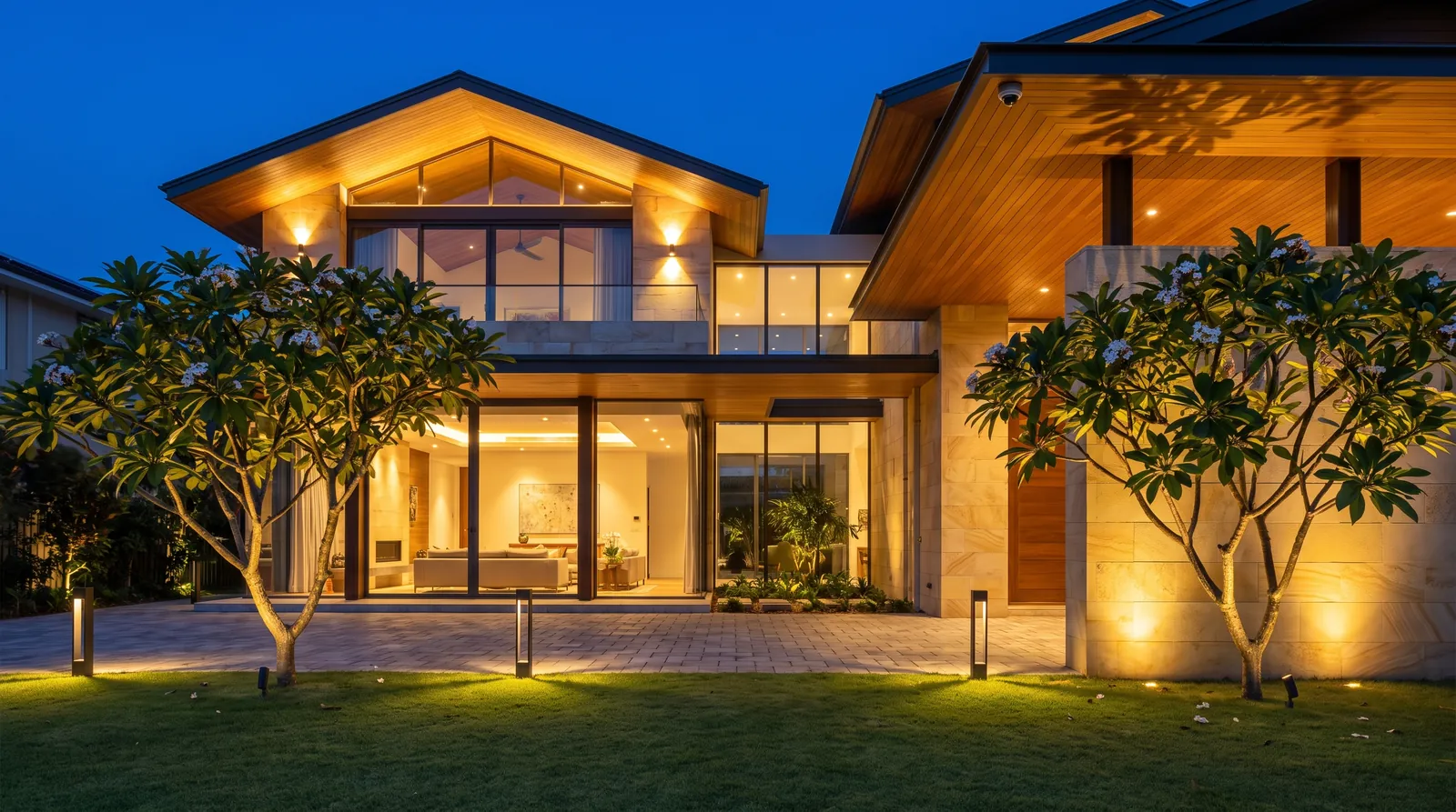 A contemporary two-storey residence at dusk, with a lit facade, uplit trees, bollards along the drive and glowing interiors.
