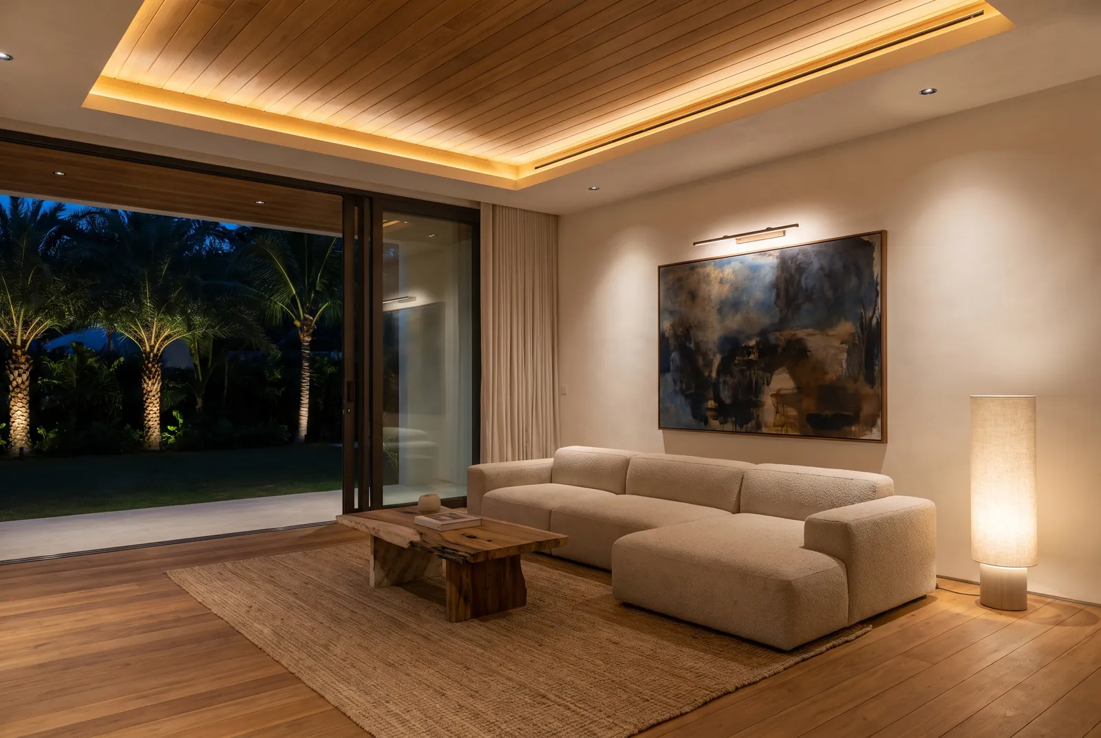 A living room in the evening lit by a warm ceiling cove, small downlights, a picture light on a large painting and a floor lamp, with uplit palms outside.