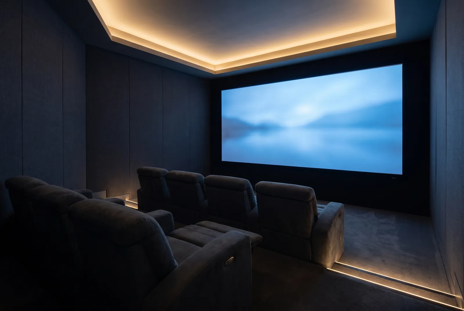 A private cinema room with dark fabric wall panels, a softly lit ceiling cove, two rows of reclining seats on a raised floor and a large screen showing a misty lake.
