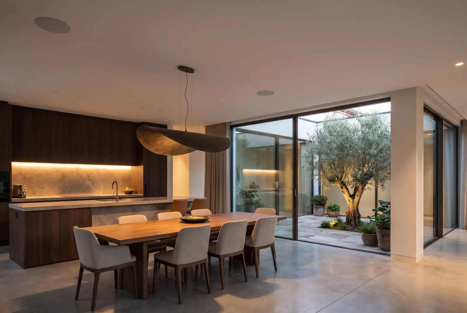 An open-plan kitchen and dining room with a warm light line under the cabinets, a sculptural pendant over the table and glass doors to a courtyard with an olive tree.