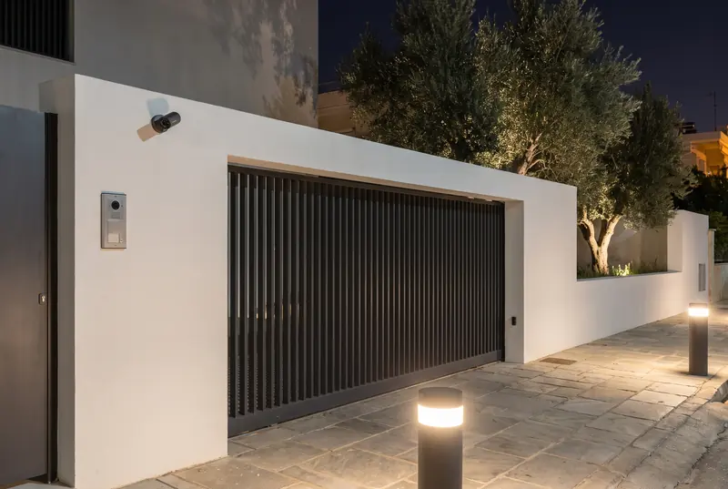 The gate of a modern residence at night, with a video door station and a discreet camera on the compound wall.